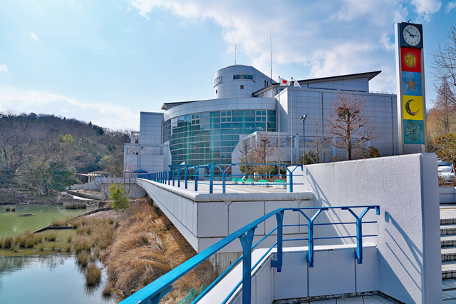 姫路科学館（アトムの館）の建物外観。メタリックなシルバーの近代建築と、太陽や月のマークが入った特徴的な時計塔。青空と池のほとりに建つ、赤穂・姫路エリアで人気の知育スポット。